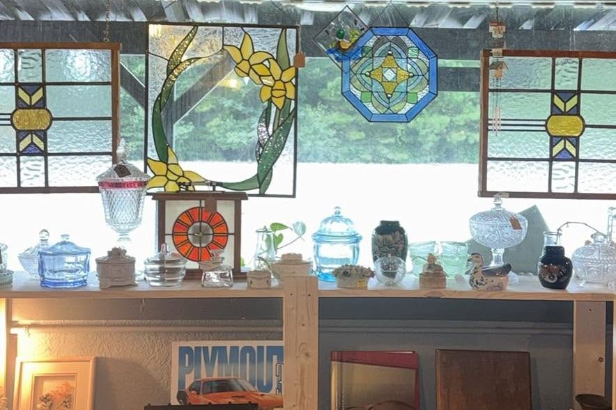 Stained-glass panels hung in a window above shelves of vintage glassware at Loblolly Gathered Goods