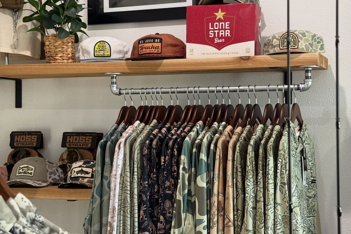 Rack of camo and print fishing shirts under a shelf of caps at Marsh House Supply Co.