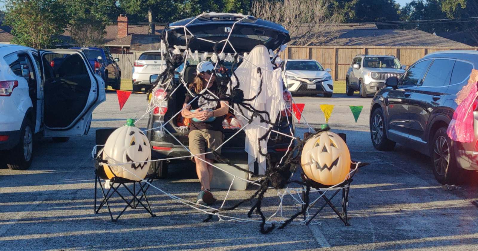 A trunk-or-treat car trunk wrapped in a giant spider web, with a ghost, a black spider and two lit jack-o'-lanterns on folding chairs