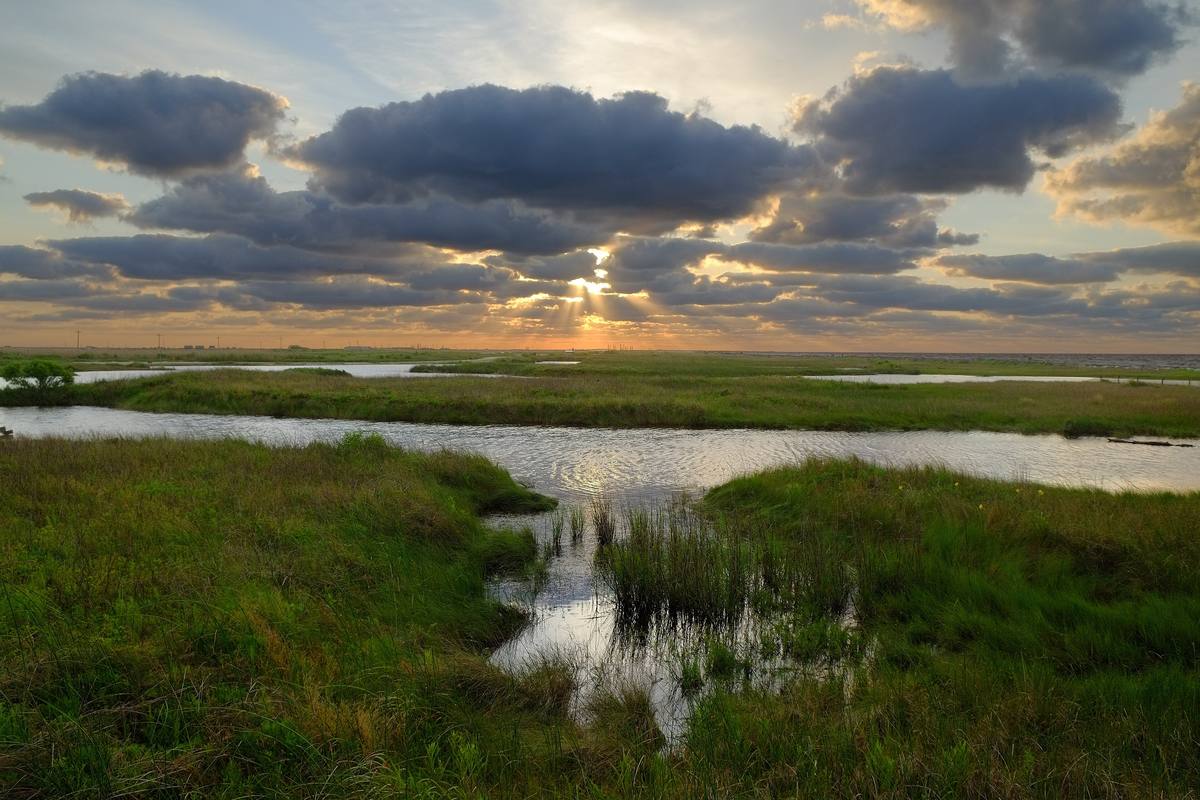 Sun breaking through clouds over salt marsh grass and open water at Sea Rim State Park