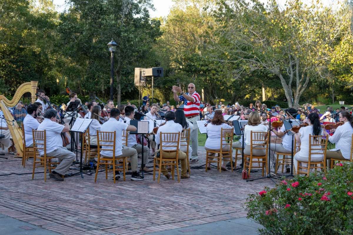 An orchestra in white shirts plays on a brick patio under the trees at the Beaumont Botanical Gardens, with a harp on the left and a crowd on the lawn behind