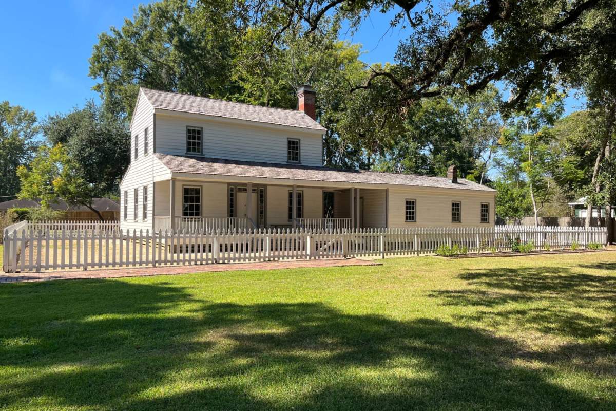 The John Jay French House in Beaumont, a white two-story wooden house with a long front porch behind a white picket fence and a wide lawn