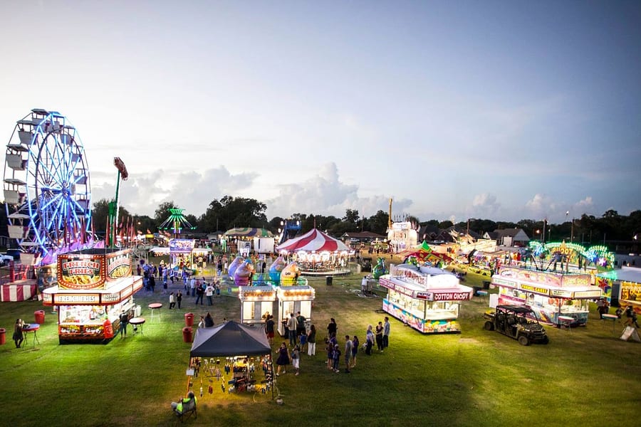The Groves Pecan Festival midway at dusk, with a lit Ferris wheel, a carousel and food trailers selling French fries and cotton candy on the grass at Lions Park