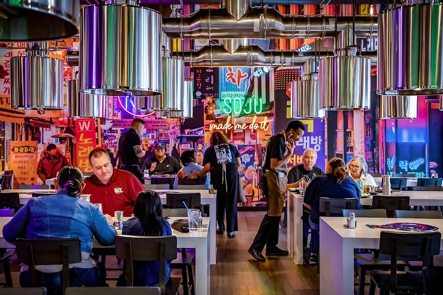 The neon-lit dining room at KO Korean Grill, Beaumont, with a steel vent hood over each table and a sign reading SOJU made me do it